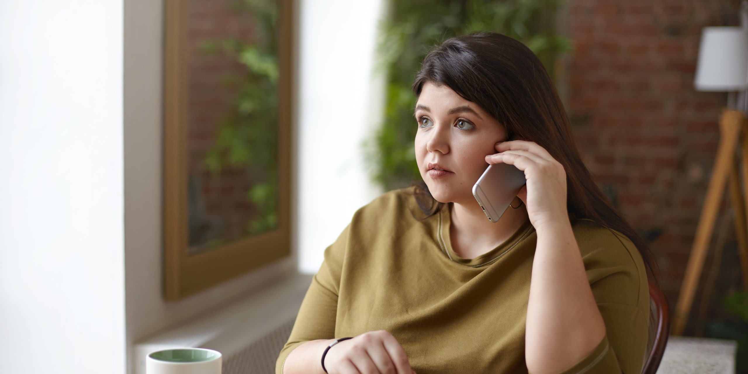 Fashionable attractive young plus size woman speaking on cell phone while drinking coffee at cafeteria with copybook lying on table in front of her. People, technology, lifestyle and communication