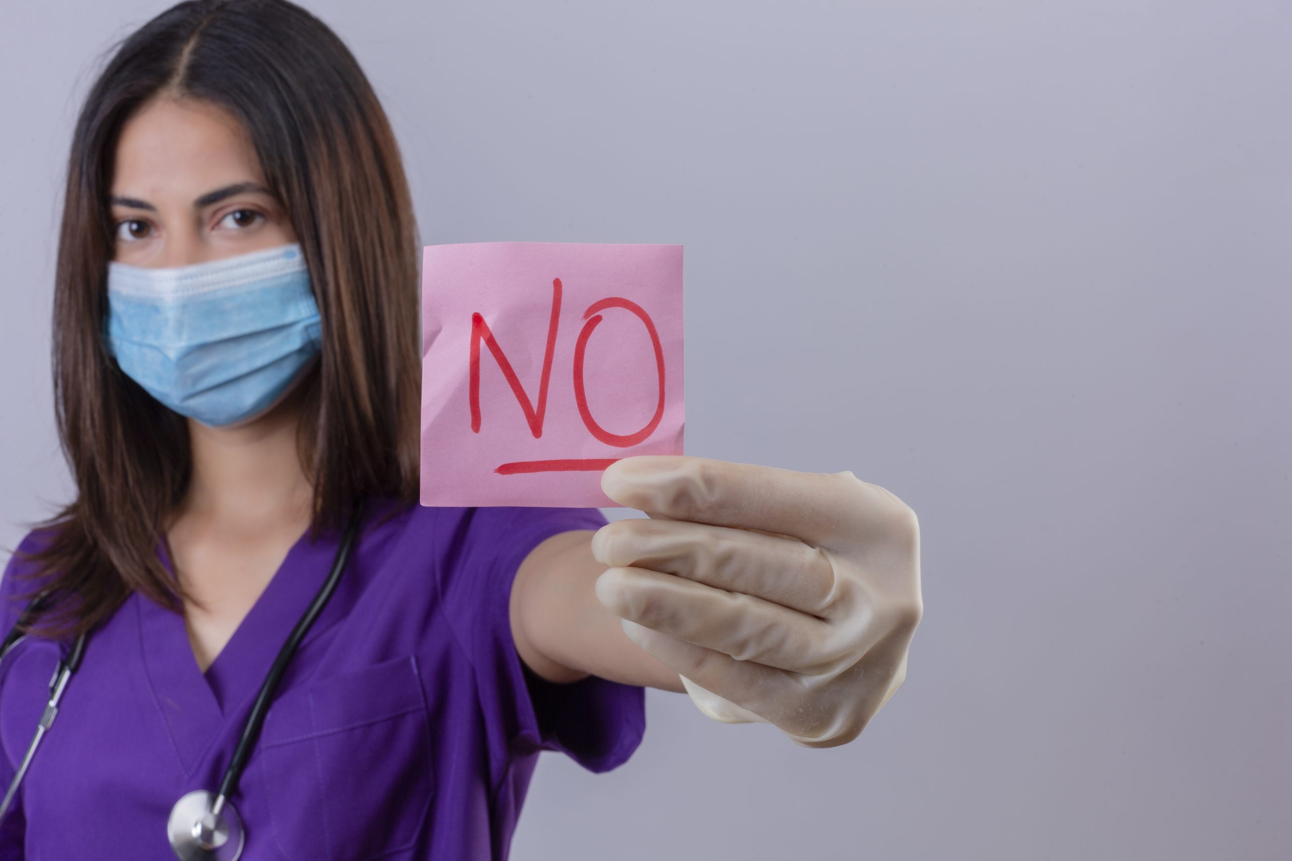 young woman nurse wearing medical uniform protective mask gloves and with stethoscope showing reminder paper with word no looking confident over white background