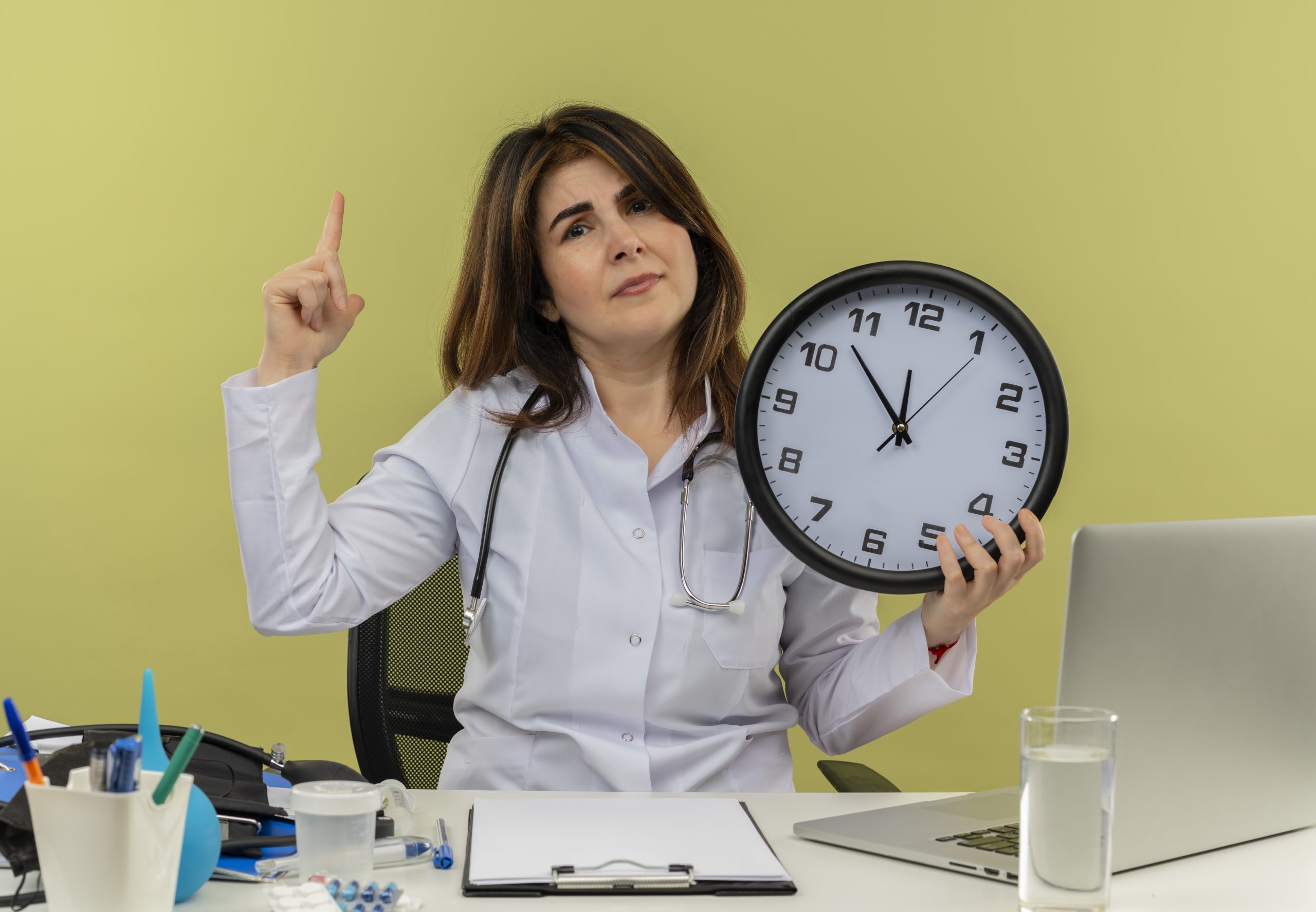 unpleased middle-aged female doctor wearing medical robe and stethoscope sitting at desk with medical tools and laptop holding clock pointing up isolated on olive green background